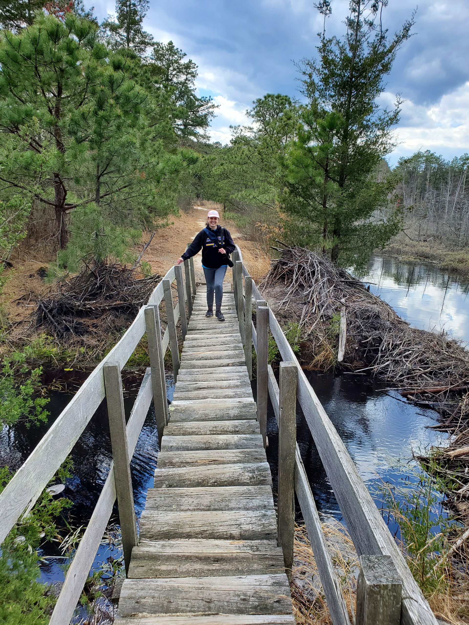 The author crossing a rough-hewn wooden bridge over water. Pine trees in the background.